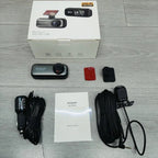 A bDashyLynk laid out on a light gray wood surface, including the retail box, a small black dash camera, mounting accessories, a car charger cable, a mounting bracket cable, a red mounting piece, and an instruction booklet.