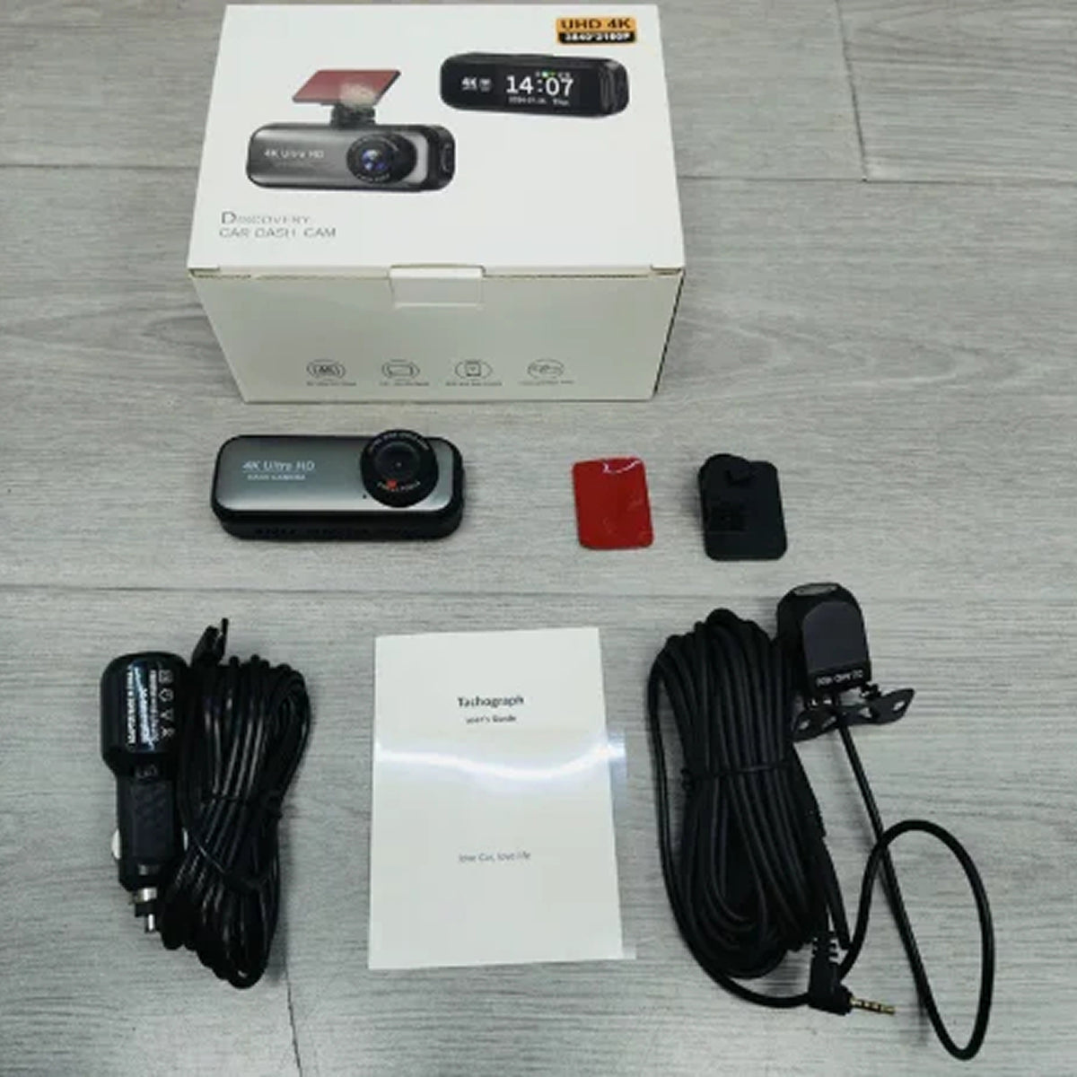 A bDashyLynk laid out on a light gray wood surface, including the retail box, a small black dash camera, mounting accessories, a car charger cable, a mounting bracket cable, a red mounting piece, and an instruction booklet.