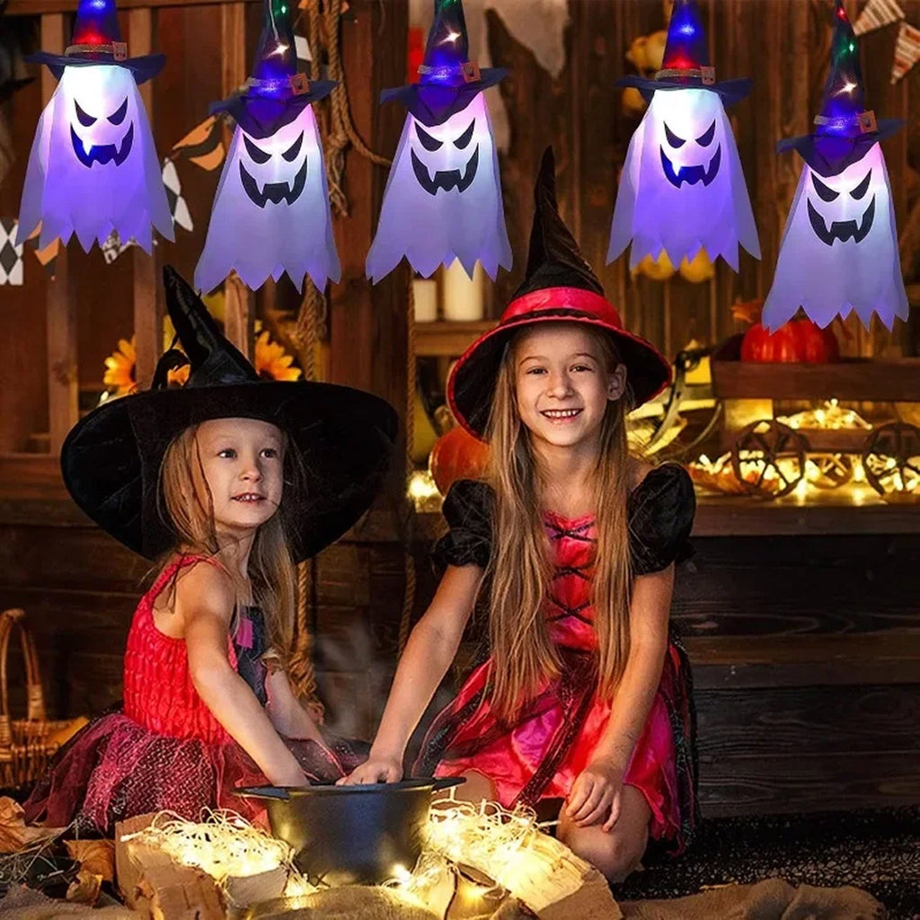 Two young girls in witch costumes sitting by a cauldron in a rustic Halloween-themed room, with glowing HexWitchi Jack O Lanterns  hanging above them.