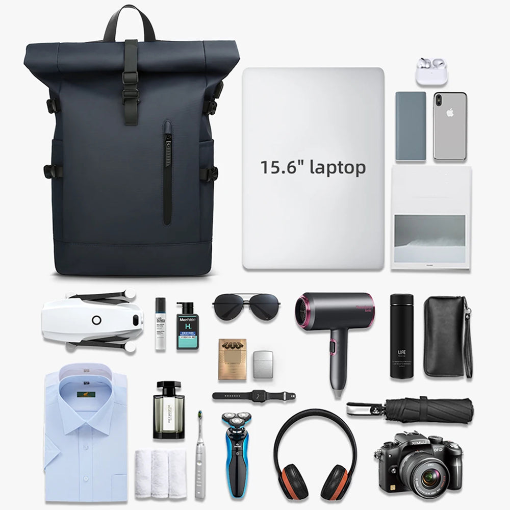 A roll-top b SafeiPack Ultimate Anti-Theft Backpack in dark navy blue is shown alongside a 15.6" laptop, wireless earbuds, sunglasses, hair dryer, camera, shirt, toiletries, and other accessories arranged on a clean white background.
